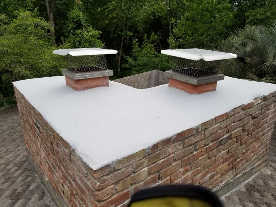 Two brick chimneys with metal caps on a white flat roof, surrounded by trees.