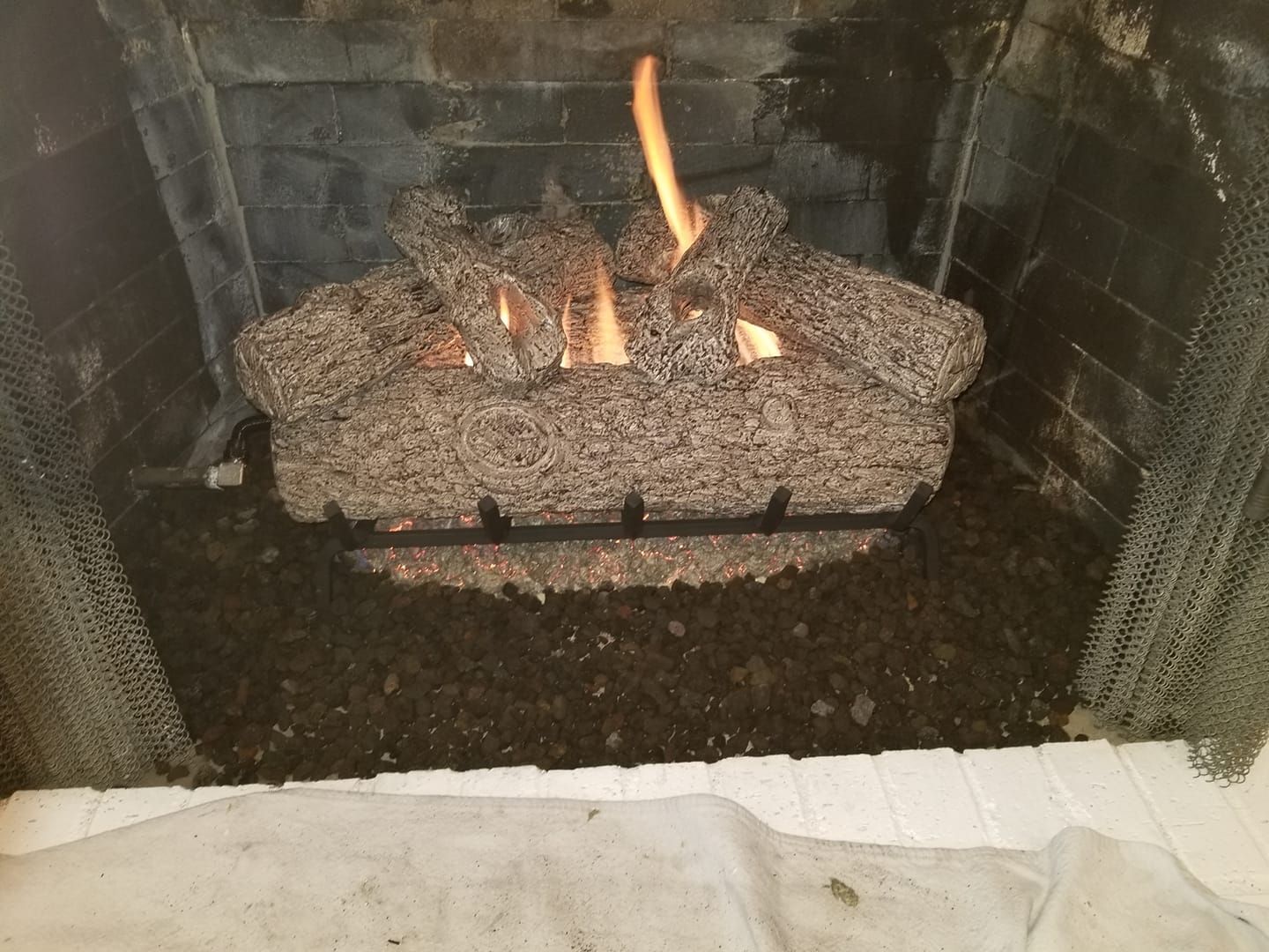 Gas fireplace with flame, gray mesh logs in a black grate, surrounded by dark granules, inside a stone hearth.