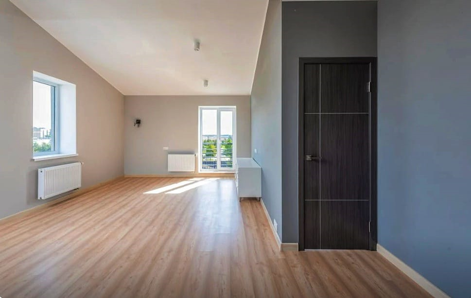 Empty room with wood floors, windows, radiators, and a dark brown door; grey and blue walls.