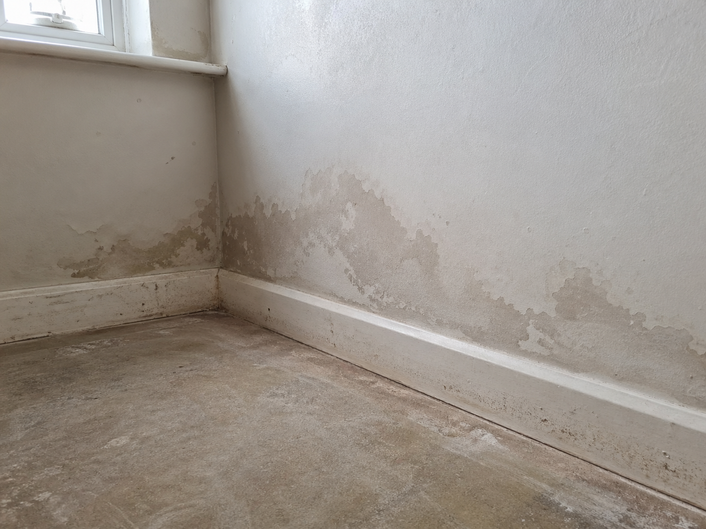 Empty corner with peeling paint and water damage on white walls and floor near a window
