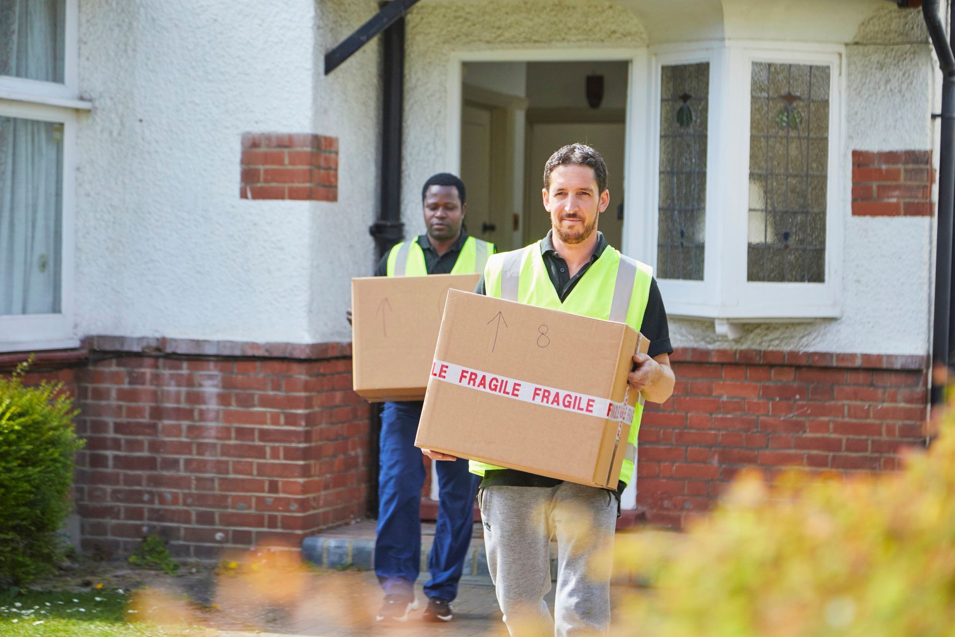 Two male movers wearing reflective vests and carrying boxes, one of whom is smiling.