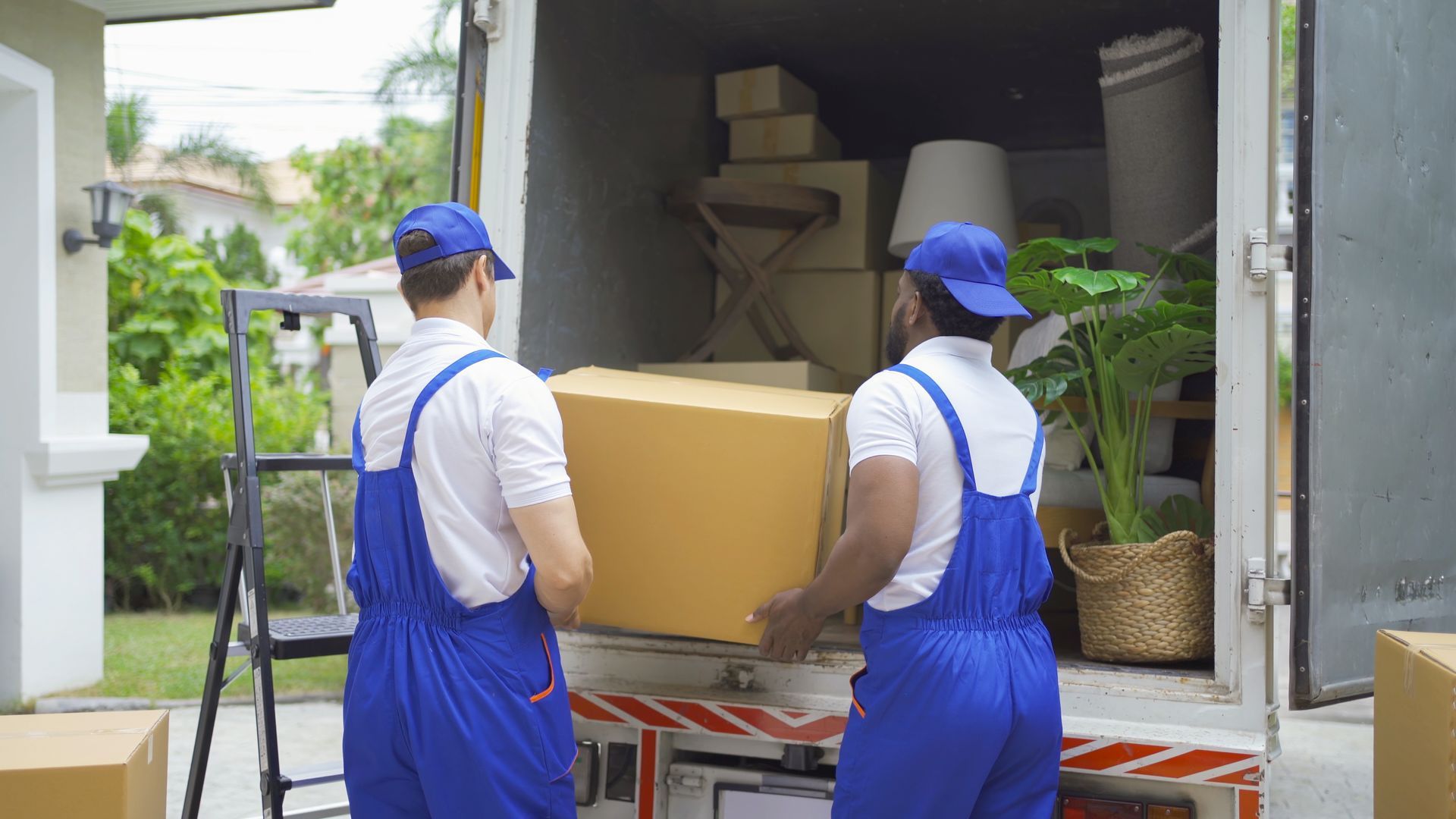 A pair of movers in work uniform lift a brown box, to place it inside a moving van.