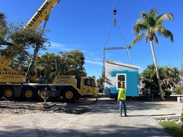 Crane lifting a small blue house while a worker stands nearby on a sunny street lined with palm trees.