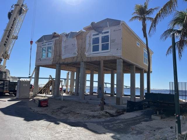 Raised house under construction on stilts near a beach, with a crane and palm trees in the background.