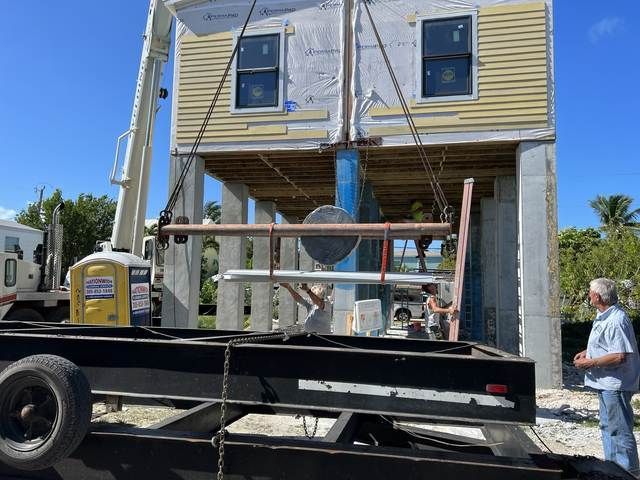 House raised on stilts by crane with workers, yellow siding and black flatbed truck in front.
