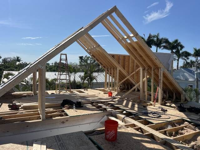 House frame under construction on a sunny lot, with wooden trusses and scattered materials around it.