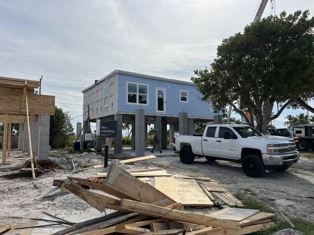 Elevated blue house under construction with a white pickup truck and building materials in the foreground.