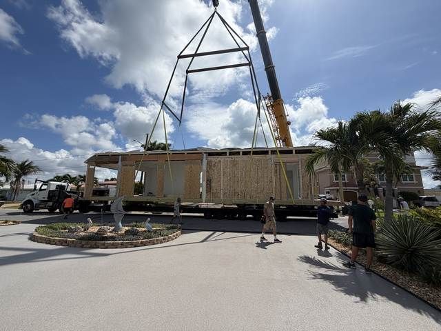 Crane lifting a large modular building onto a flatbed truck on a sunny street, with workers nearby.