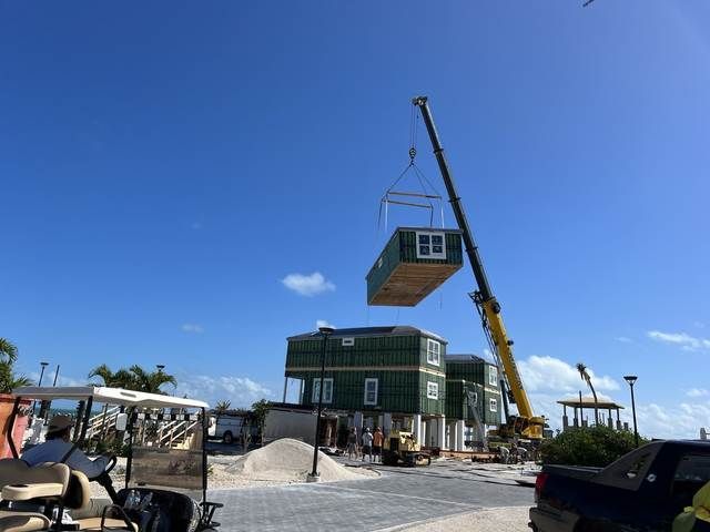 Crane lifting a tiny house onto a site with a green building under a clear blue sky.