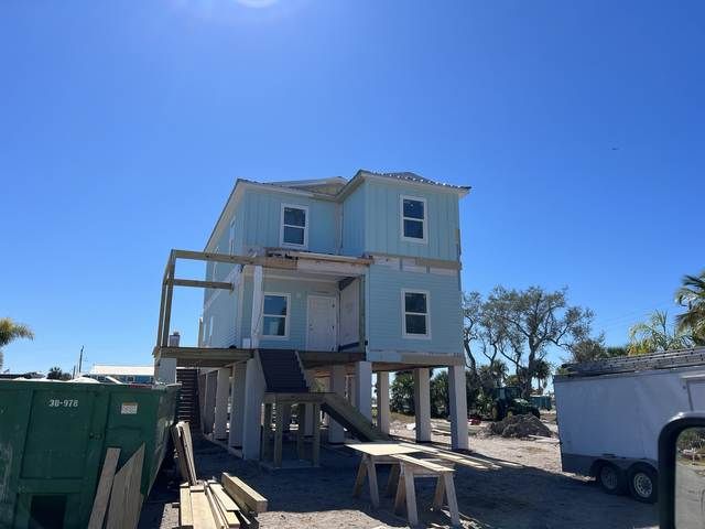 Two-story house under construction with exterior stairs, blue siding, and construction vehicles nearby.