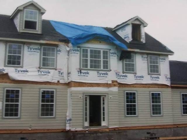 House under construction with Tyvek wrap, blue tarp on roof, and front door open.