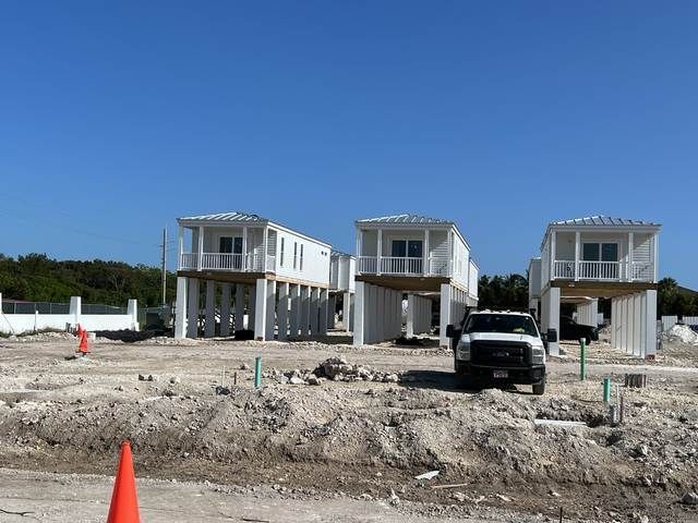 Three elevated houses under construction on a dirt lot with a parked black truck and orange cones