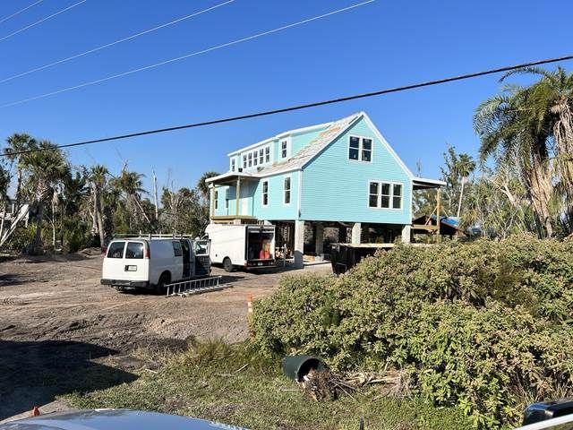 Light blue raised house on stilts with two vans parked on a sandy lot under a clear blue sky.