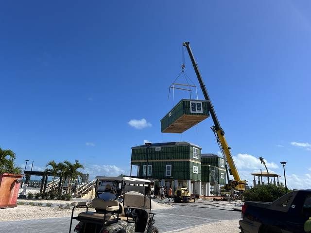 Crane lowering a green house module onto a two-story building under construction near the coast.