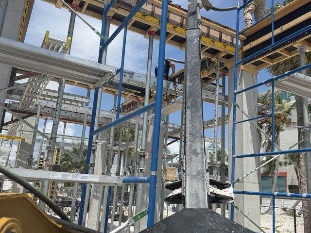 Steel framework of a building under construction, with blue scaffolding and palm trees in the background.