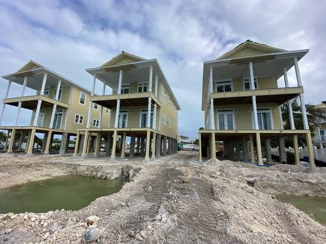 Three stilt houses under construction on a sandy waterfront, with a small pond in front.