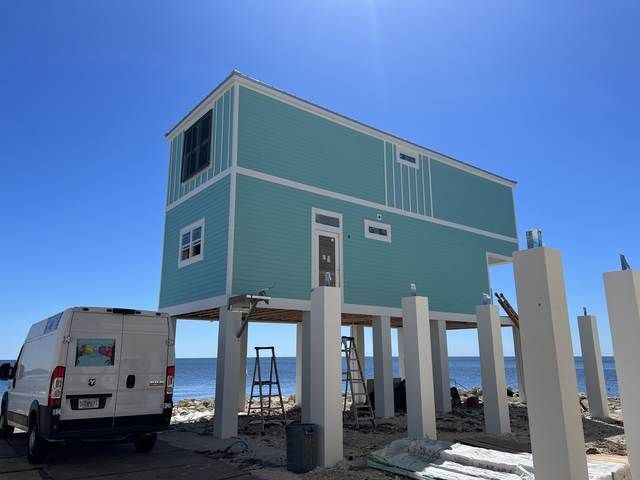 Elevated turquoise house on stilts by the beach, with a white van parked beside it under a clear blue sky.