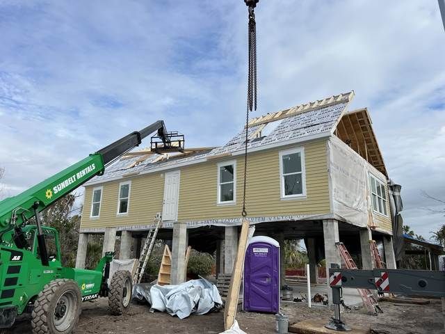 House under construction with green crane lifting materials beside a two-story home on stilts.