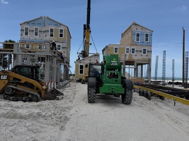Construction site with two houses on stilts, crane, excavator, and green lift on a sandy beach.