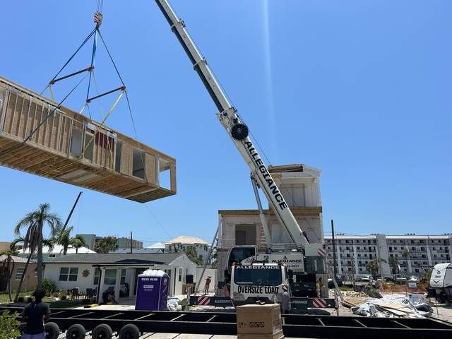 Crane lifting a long wooden structure at a sunny construction site near buildings and a purple porta-potty.