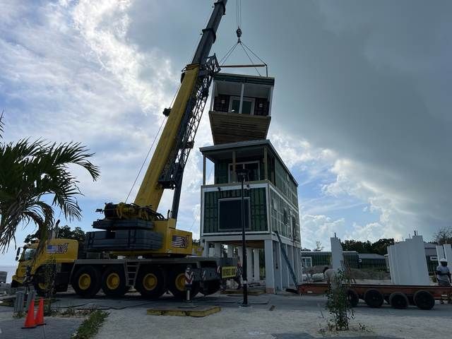 Crane lifting a small tower-like building module at a construction site under a cloudy sky.