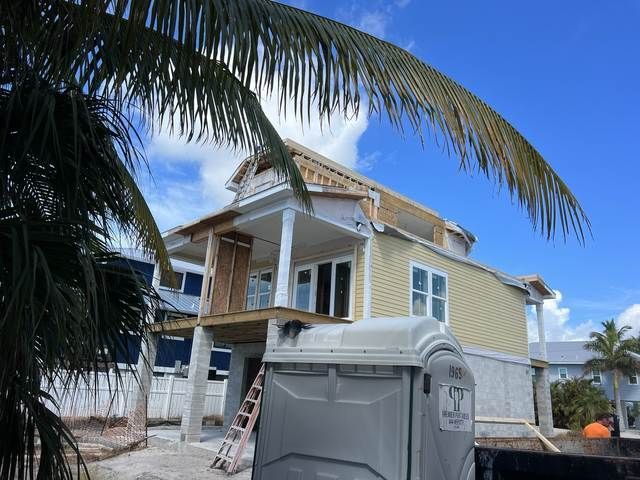 Two-story yellow house under construction with a porch and palm fronds overhead, next to a gray dumpster.