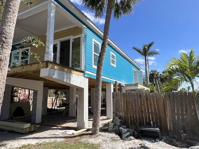 Raised turquoise beach house with white trim, palm trees, and a fenced yard under a bright blue sky.