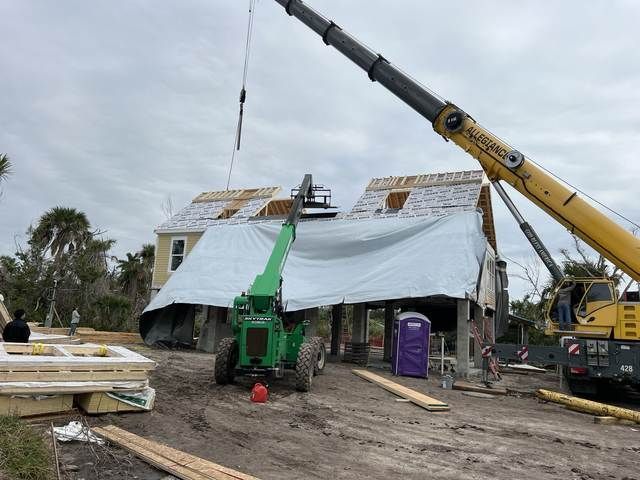 Construction site with two cranes lifting materials to a roof under repair, tarp-covered house in the center.