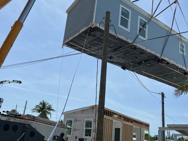 Blue house being lifted by a crane above a small building on a construction site.