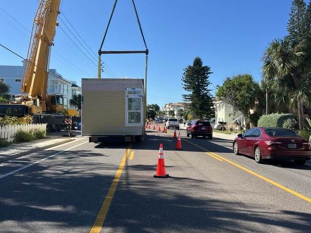 Crane lowering a small prefab building onto a street, with cones and cars nearby.