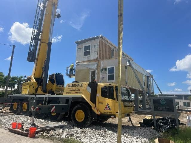 Construction crane lifting a beige house module on a truck under a blue sky.