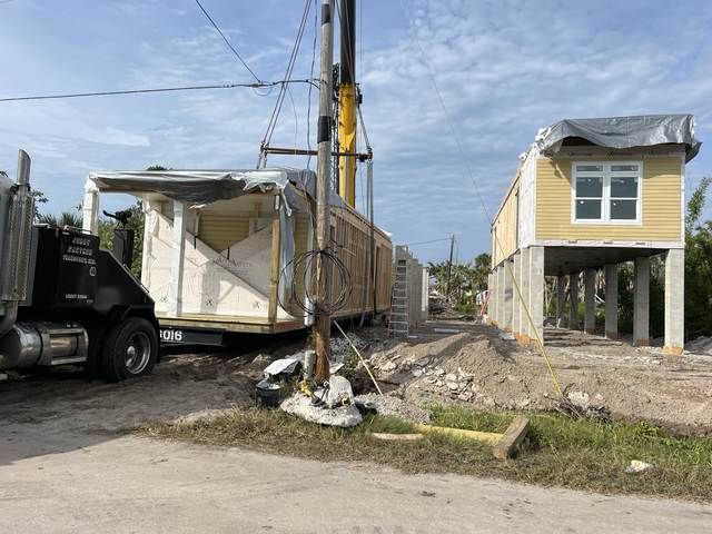 House being lifted by a crane beside a truck in a debris-strewn lot, with another house on stilts nearby.