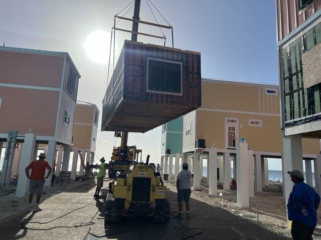 Crane lifting a modular house unit between buildings on a sunny street, with workers nearby.