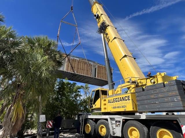 Yellow crane lifting a wooden deck over a truck beside palm trees under a blue sky.
