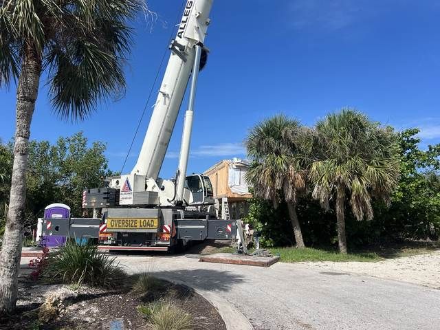 Large crane truck on a dirt road beside palm trees under a blue sky.