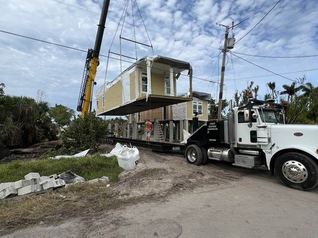 White truck hauling a modular house on a flatbed along a roadside with a crane lifting nearby.