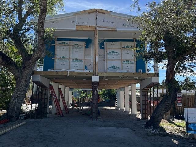 Elevated house under construction on stilts, with trees and a dirt yard in front.