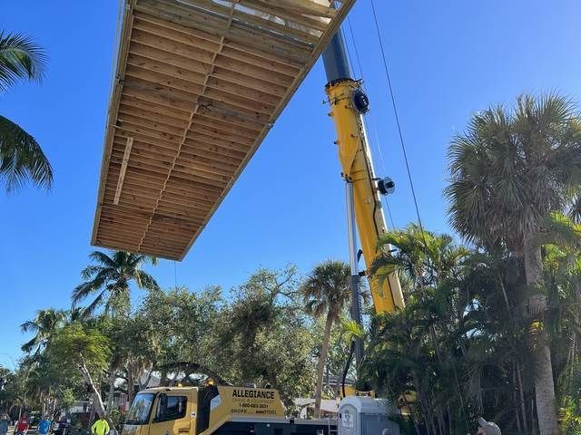 Construction crane lifting a large wooden platform above palm trees on a sunny day.