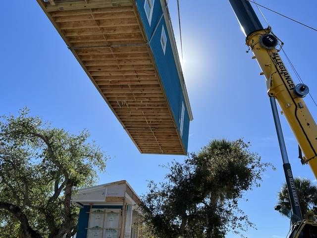Bridge section being lifted by a crane over a house and trees under a blue sky.