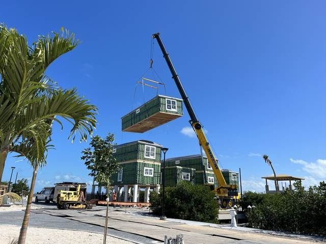 Crane lifting a green shipping container onto a stacked container structure under a clear blue sky.