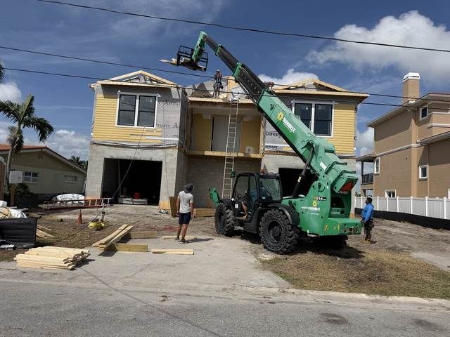 Workers use a green crane to lift materials at a yellow house under construction, with debris in the driveway.