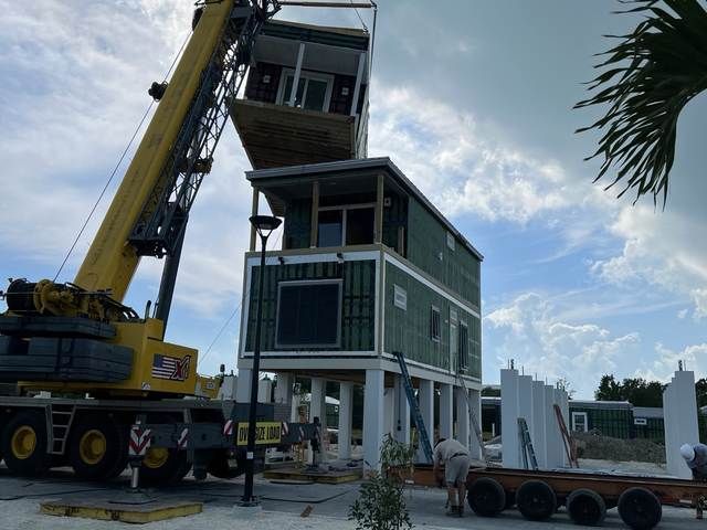 Crane lifting a small house onto a raised foundation at a construction site.