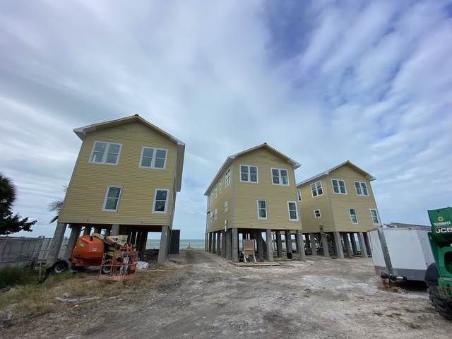 Three yellow houses on stilts under construction on a gravel lot beneath a cloudy sky.