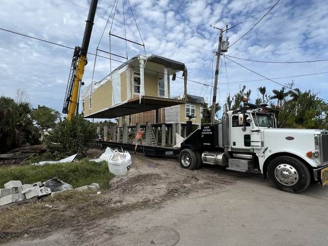 Truck hauling a modular house on a road with a crane lifting it in a tropical area.