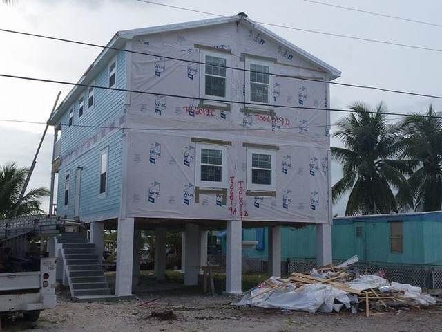 Elevated white house under construction with scattered debris and palm trees nearby.