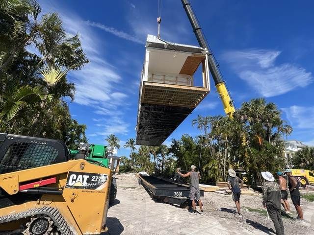 Crane lifting a large rectangular module while workers guide it on a sunny street with palm trees and trucks.