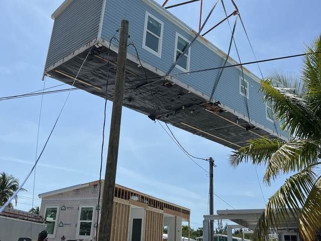 Blue house being lifted by crane over a street, with a palm tree and utility poles below.