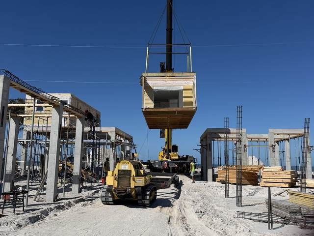 Construction site with a crane lifting a yellow modular unit over workers and steel frames under a blue sky.