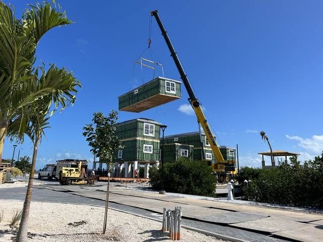 Crane lifting a green modular building unit at a construction site under a clear blue sky.