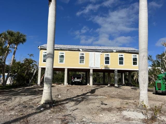 Yellow house on stilts under construction, framed by tall white columns and palm trees.